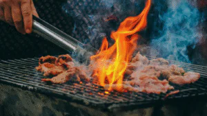 Människa med grilltång som vänder köttbitar på en grill